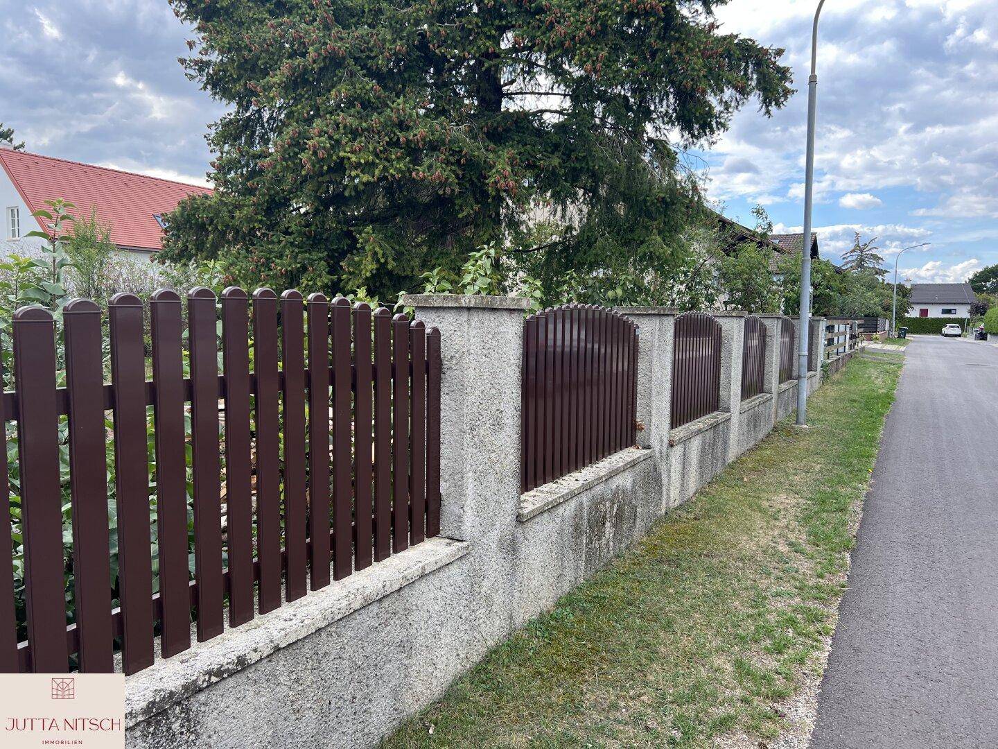 Langer Holzzaun mit Betonfundament entlang einer ruhigen Wohnstraße, umgeben von Grün.