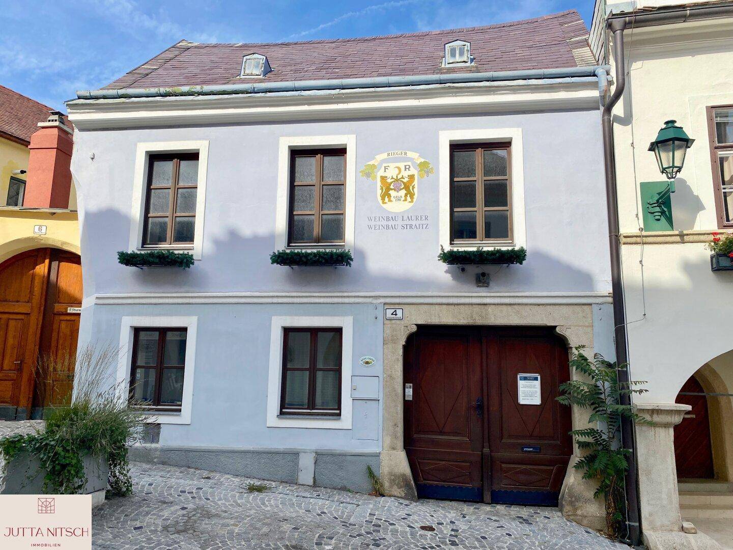 Historisches Gebäude mit hellblauer Fassade, braunen Fenstern und einem großen Holztor an einer gepflasterten Straße.