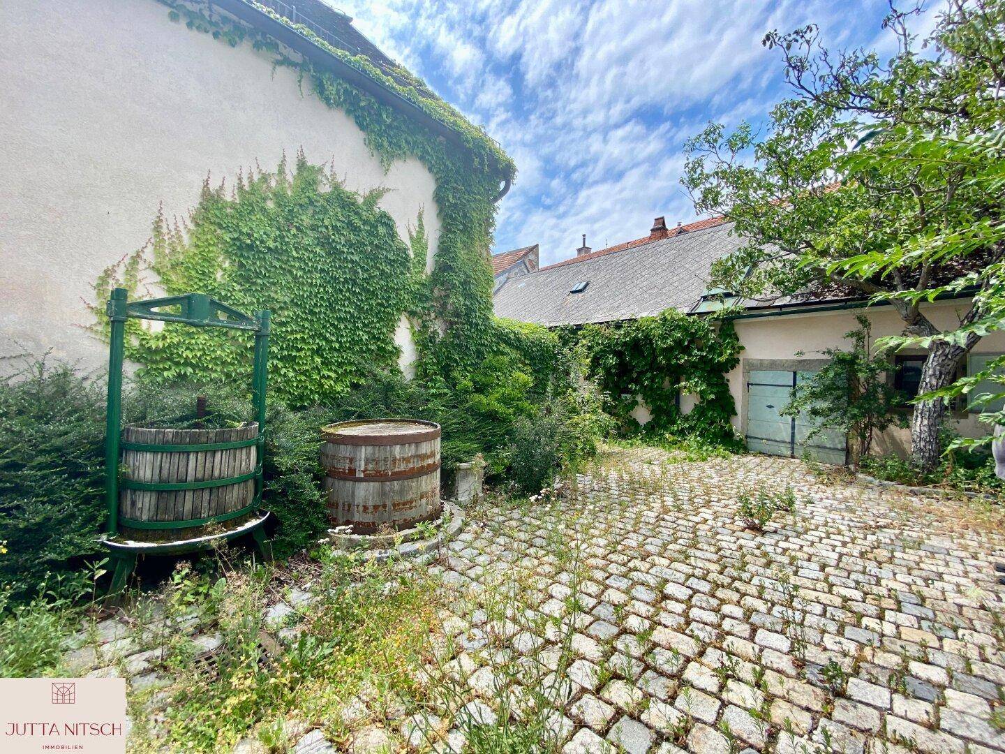 Verwilderter Innenhof mit Kopfsteinpflaster, bewachsenen Wänden und rustikalen Holzfässern.