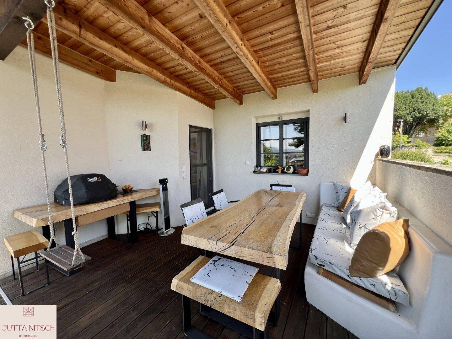 Überdachte Terrasse mit Holzbalkendecke, rustikalem Esstisch und integriertem Grillbereich.