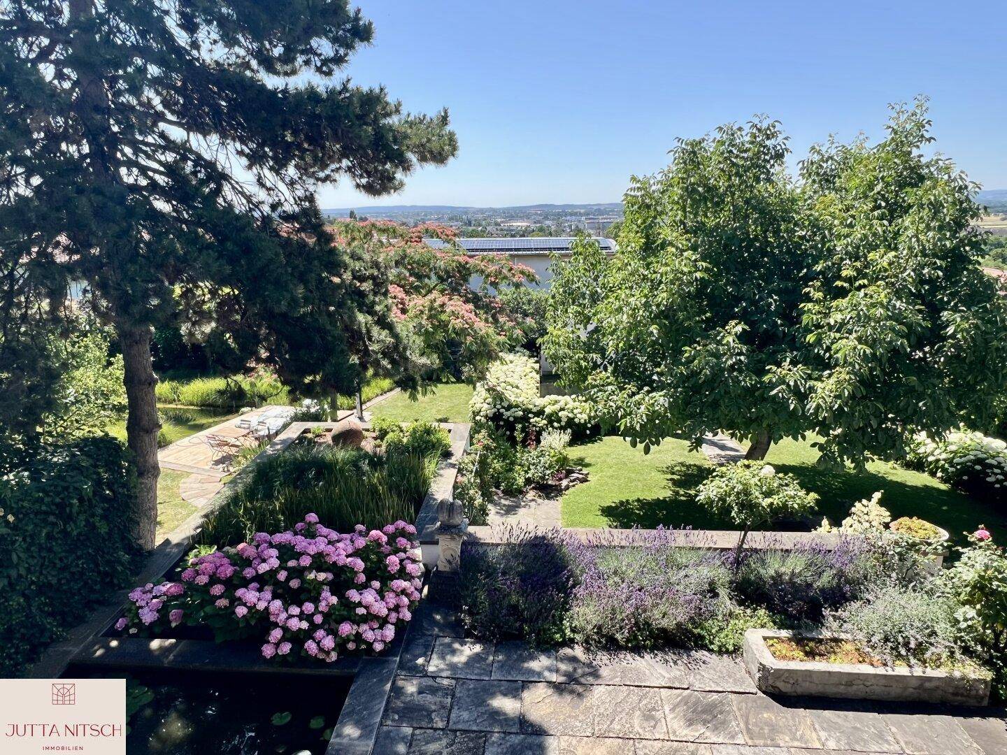 Terrassierter Garten mit Teich, blühenden Pflanzen und einem weiten Blick über die Landschaft.
