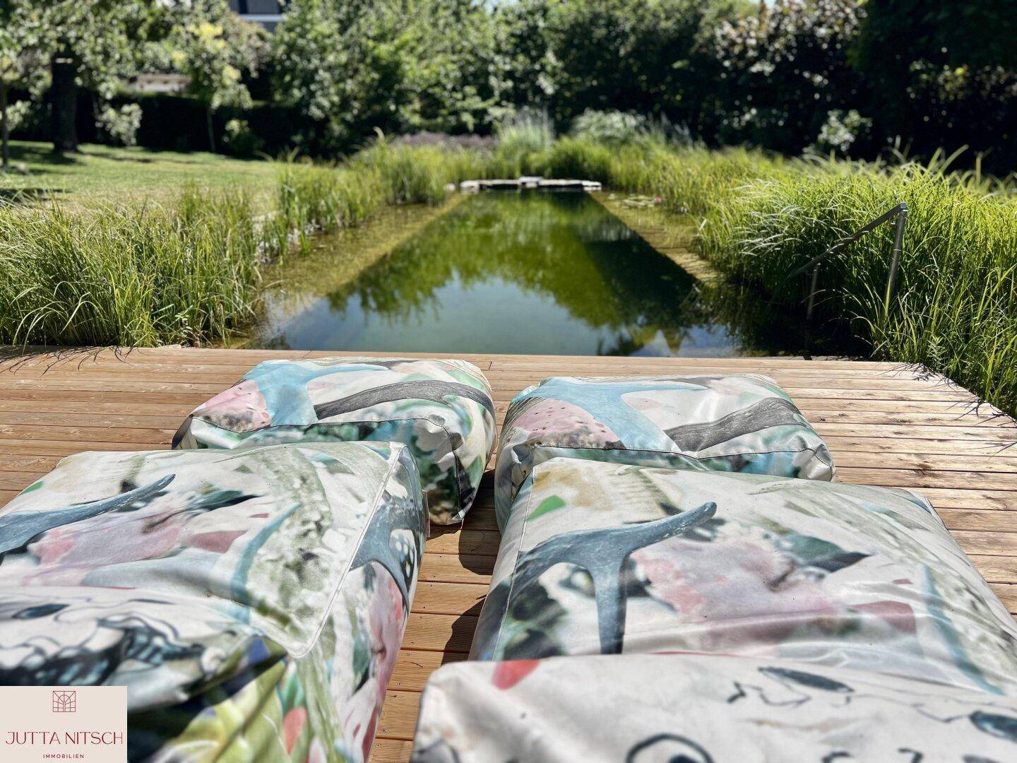Natürlicher Schwimmteich mit Holzdeck und bequemen Sitzkissen in einer grünen Gartenlandschaft.