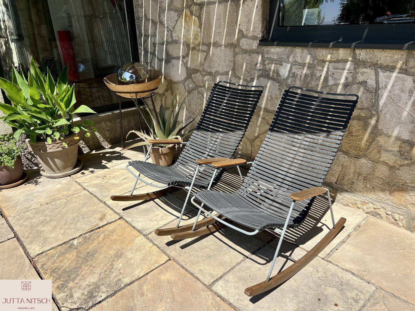 Sonnige Terrasse mit zwei modernen Schaukelstühlen und Topfpflanzen vor einer Steinwand.