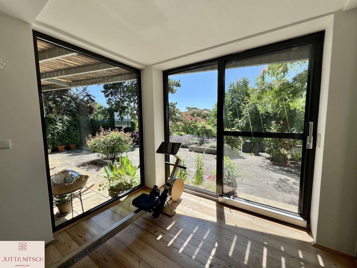 Heller Fitnessraum mit großen Fenstern und direktem Zugang zur Terrasse und zum Garten.