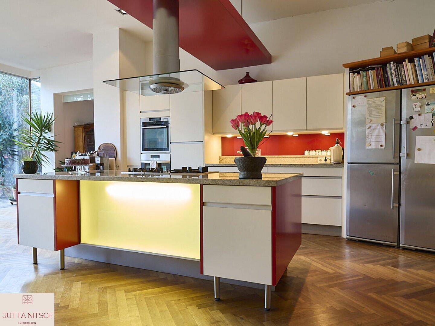 Moderne Küche mit roter Kücheninsel, integrierten Geräten und Blick in den Garten durch große Fenster.