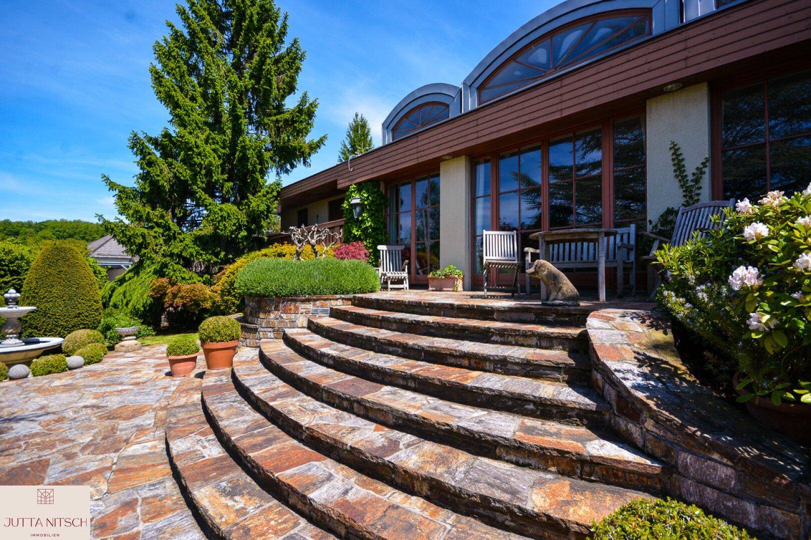 Elegante Steintreppe führt zu einer Terrasse mit Gartenmöbeln und üppiger Bepflanzung.
