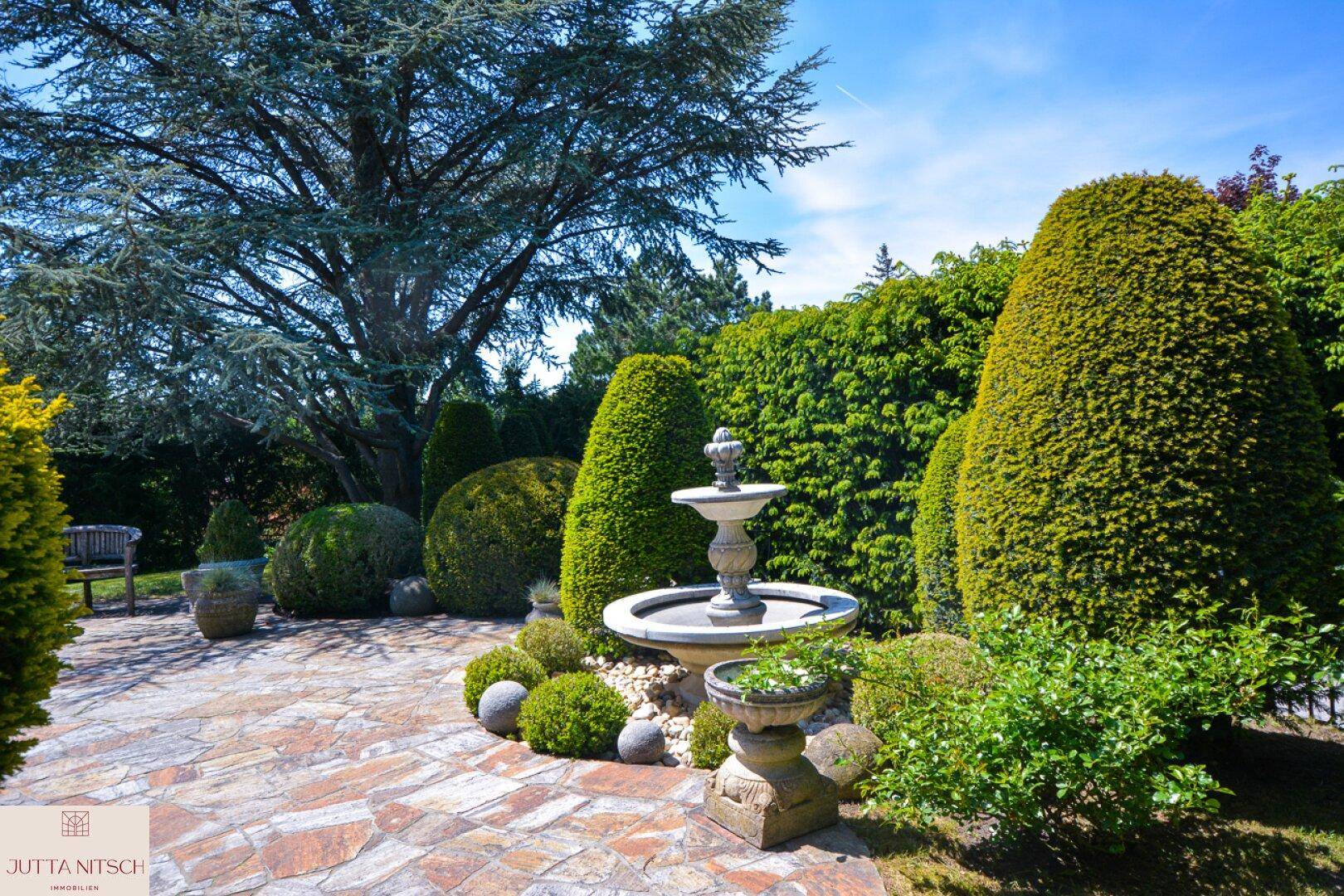 Gepflegter Gartenbereich mit dekorativem Brunnen, Steinterrasse und vielfältiger Bepflanzung.