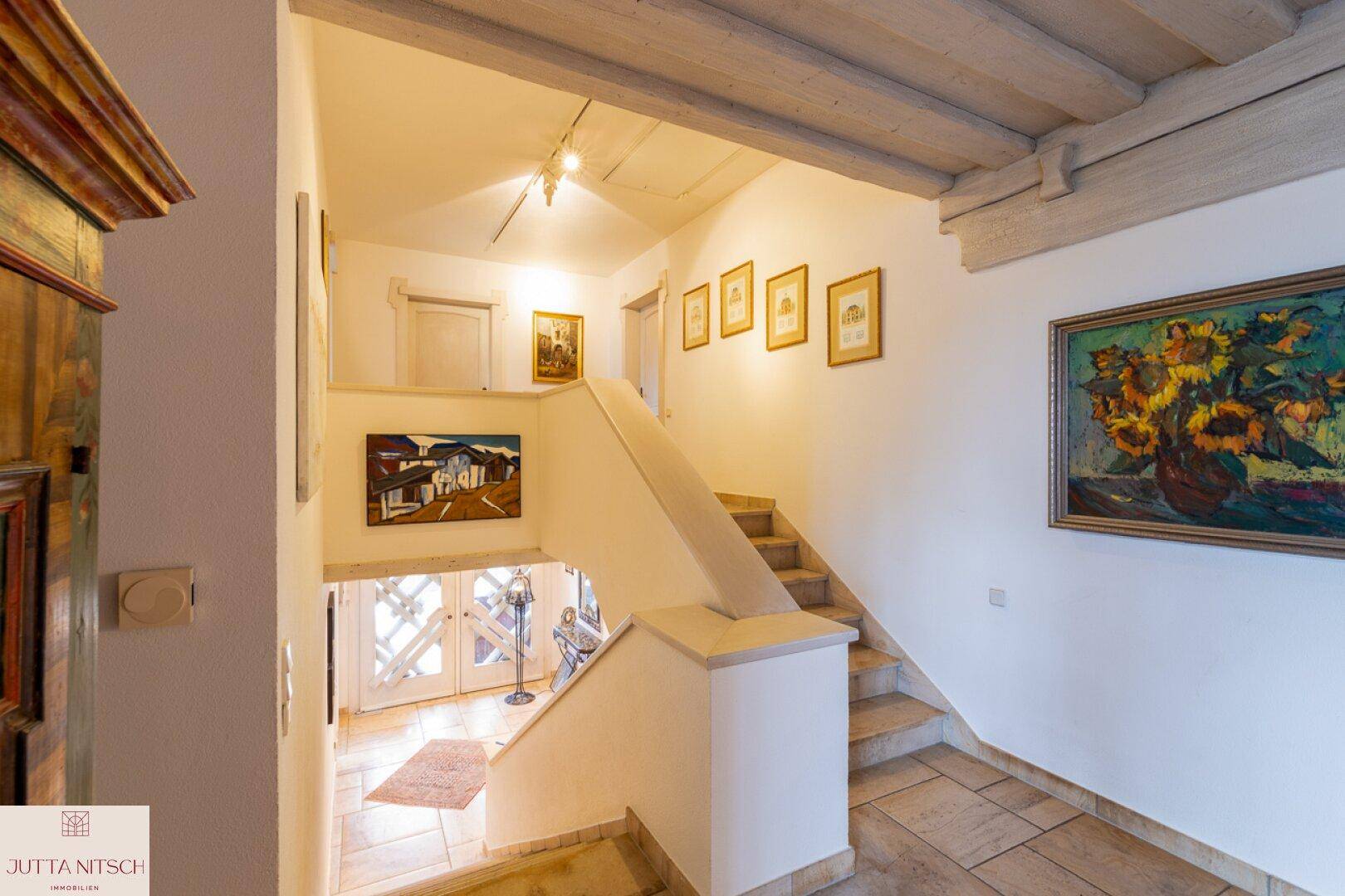 Treppenhaus mit Holzbalkendecke, Fliesenboden und Kunstwerken an den Wänden.
