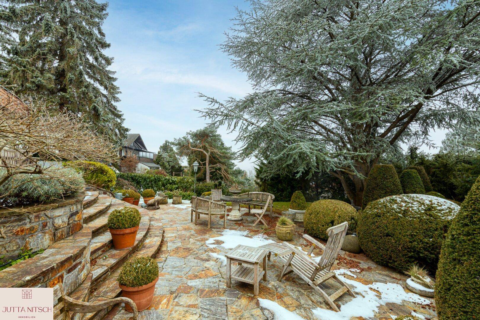 Winterliche Steinterrasse mit Gartenmöbeln, umgeben von schneebedeckten Bäumen und Sträuchern.