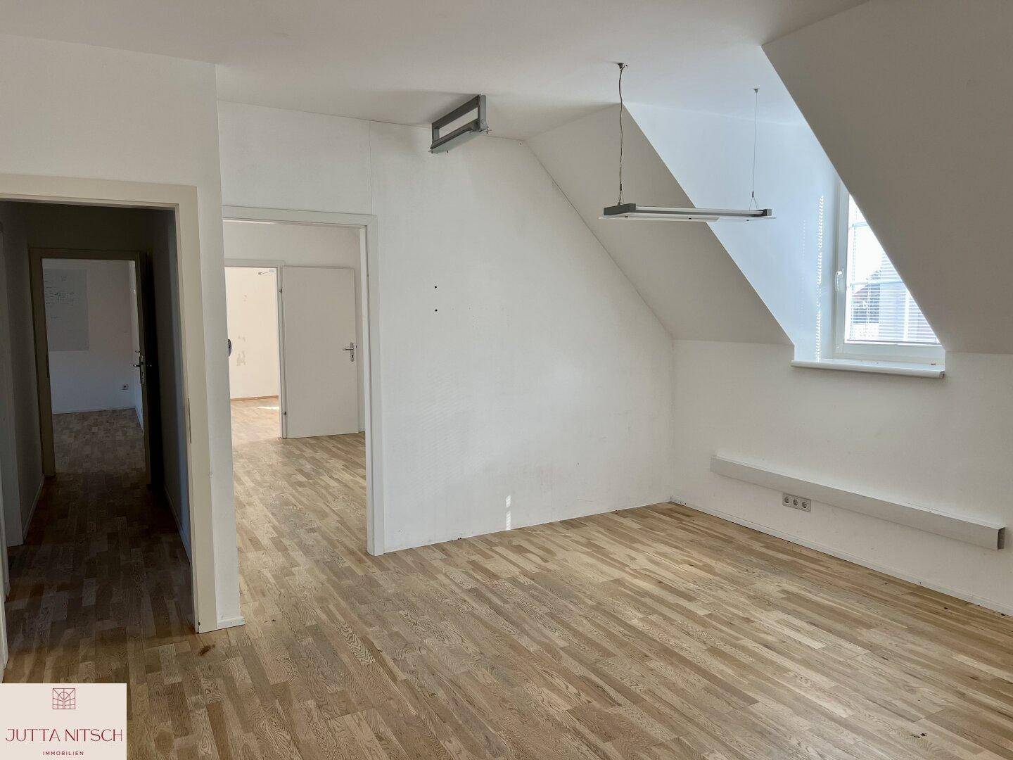 Heller Raum im Dachgeschoss mit Holzboden, schrägen Wänden und einem Fenster mit Blick nach draußen.