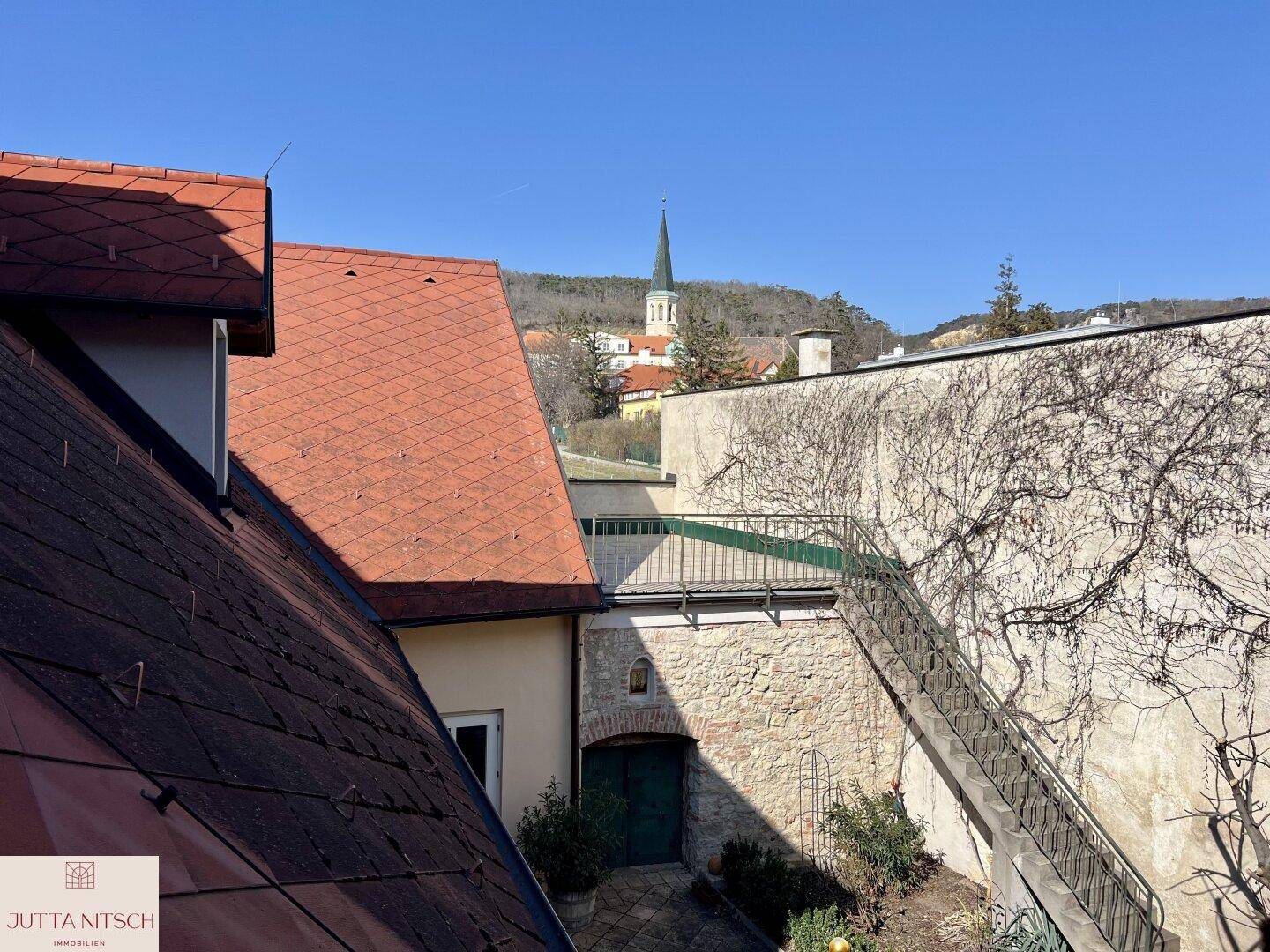 Panoramablick über rote Ziegeldächer auf eine Kirche und die umliegende Landschaft unter blauem Himmel.