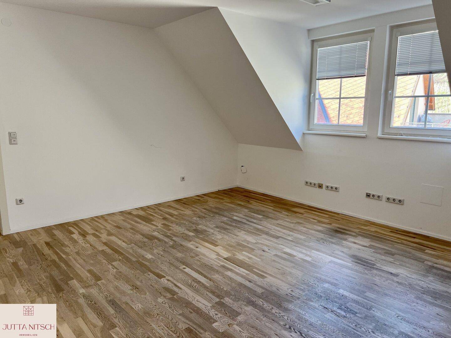 Geräumiger Dachgeschossraum mit Holzboden, schrägen Wänden und zwei Fenstern für viel Tageslicht.