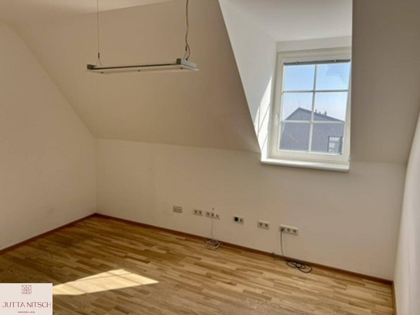 Heller Dachgeschossraum mit Holzboden, schräger Decke und einem Fenster mit Blick auf Dächer.