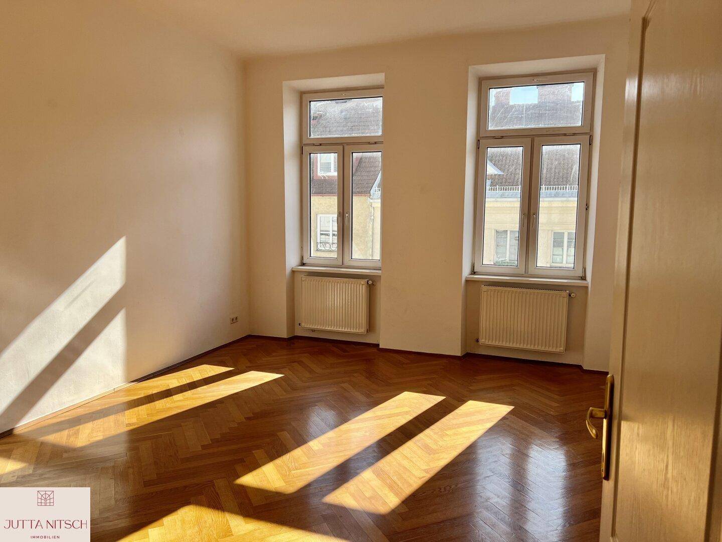 Lichtdurchfluteter Raum mit Fischgrätparkett und großen Fenstern, ideal als Wohn- oder Schlafzimmer.