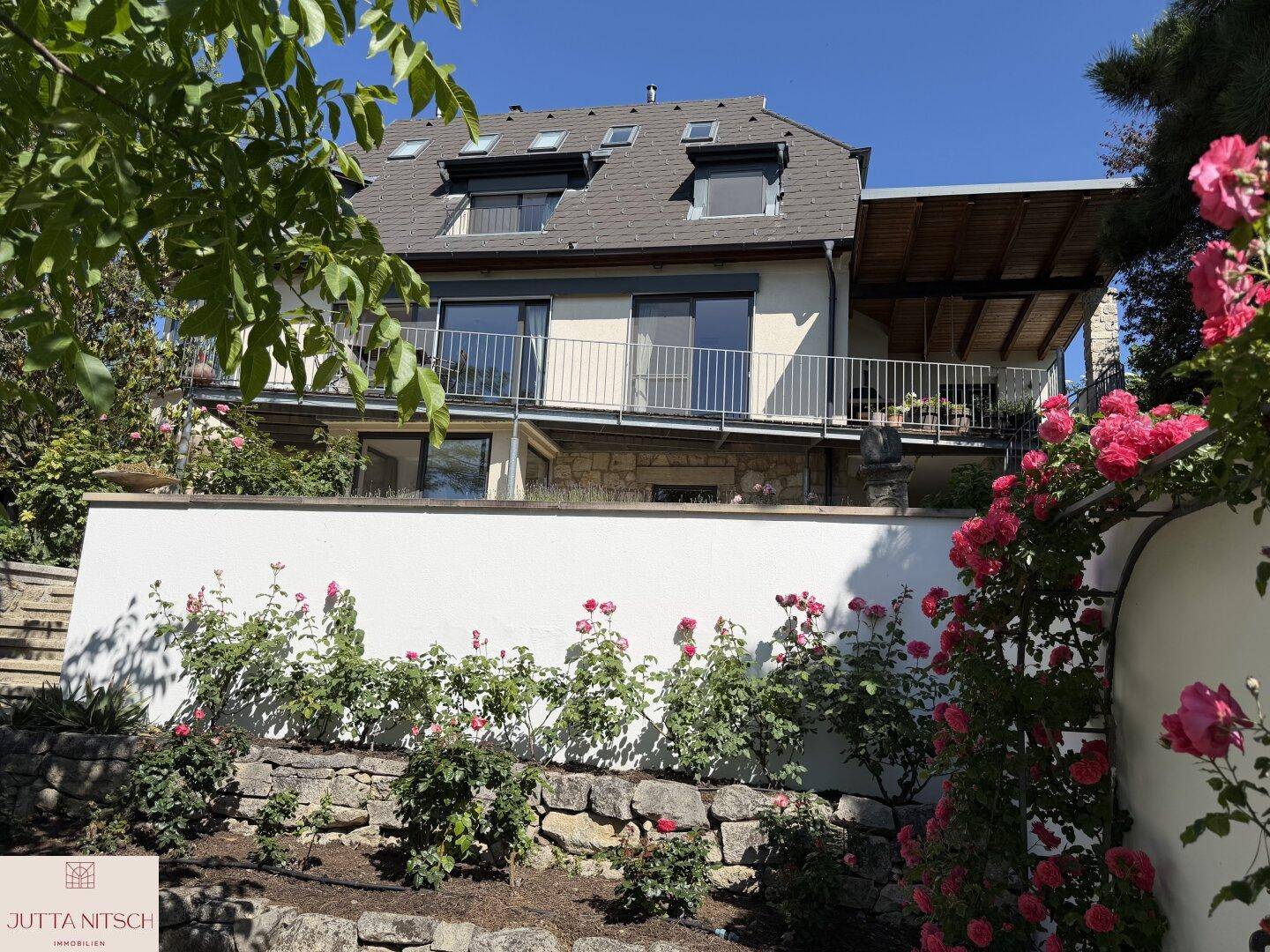 Hausfassade von der unteren Gartenebene aus gesehen, mit blühenden Rosen und einer Steinmauer.