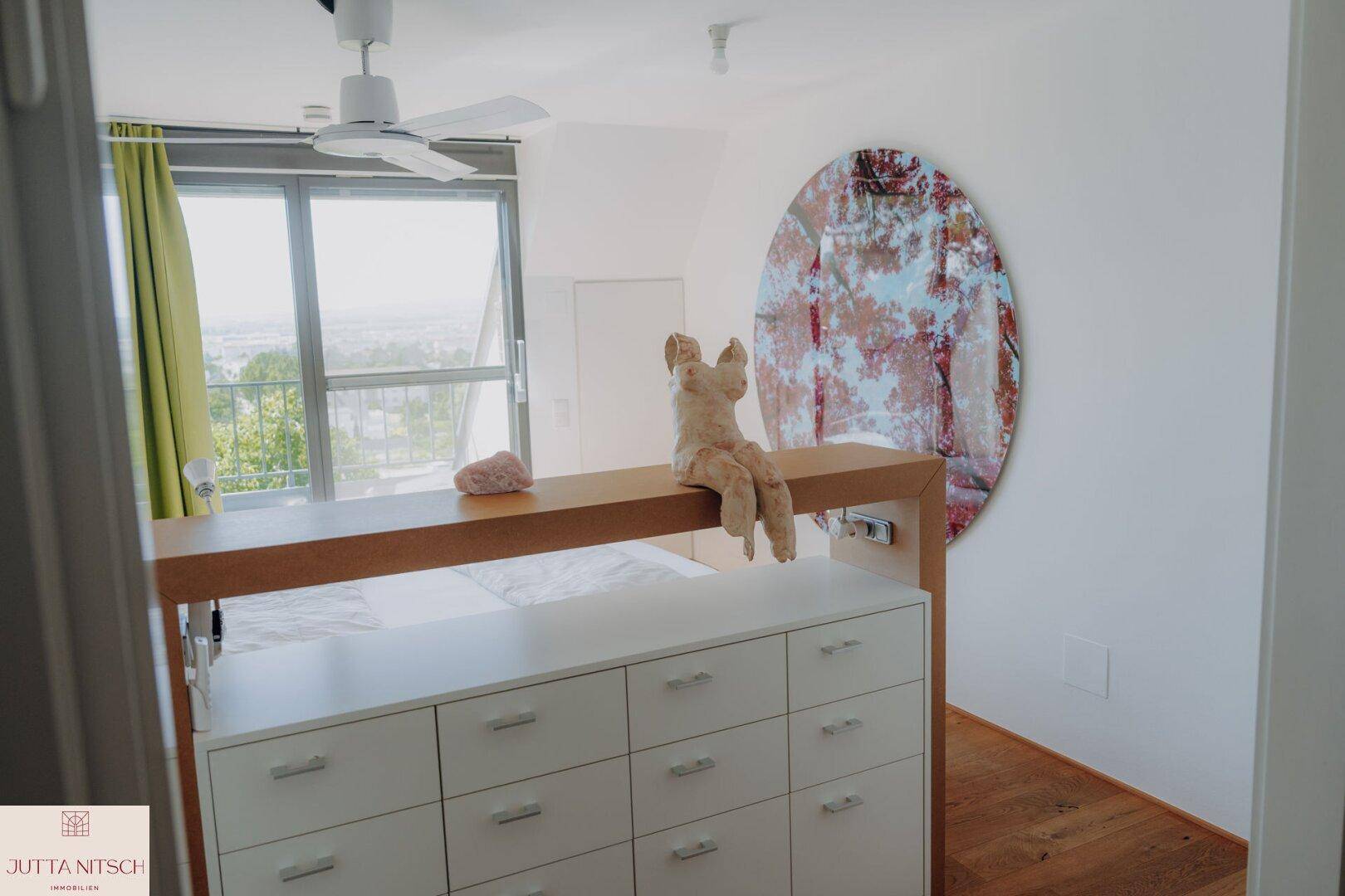 Helles Schlafzimmer mit Bett, Kommode, Deckenventilator und großen Fenstern mit Ausblick.