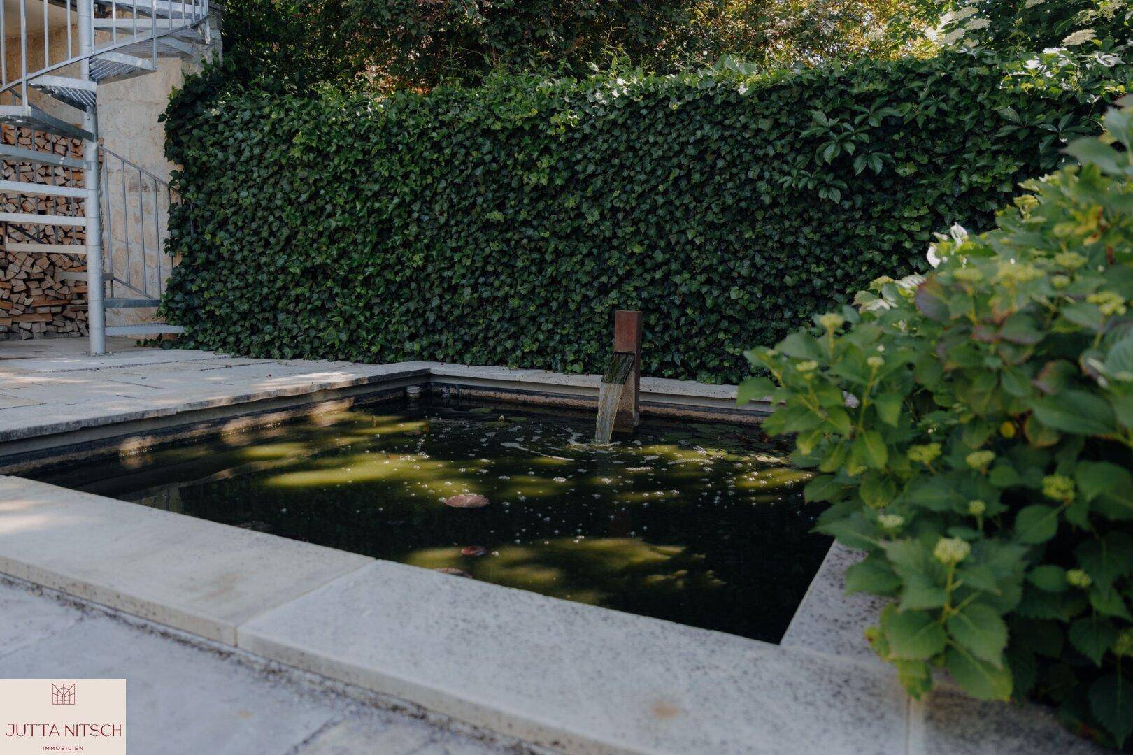 Detailansicht des Gartenteichs mit einer grünen Hecke im Hintergrund und einer Wendeltreppe.