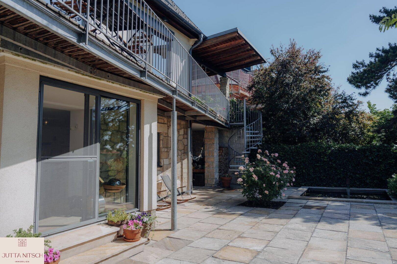 Gepflasterte Terrasse im Erdgeschoss mit großen Glasschiebetüren und einer Wendeltreppe zum oberen Balkon.