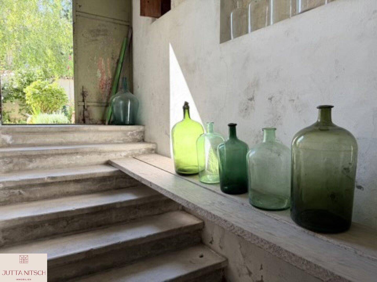 Steintreppe führt zu einem hellen Außenbereich, gesäumt von großen grünen Glasflaschen als Dekoration.