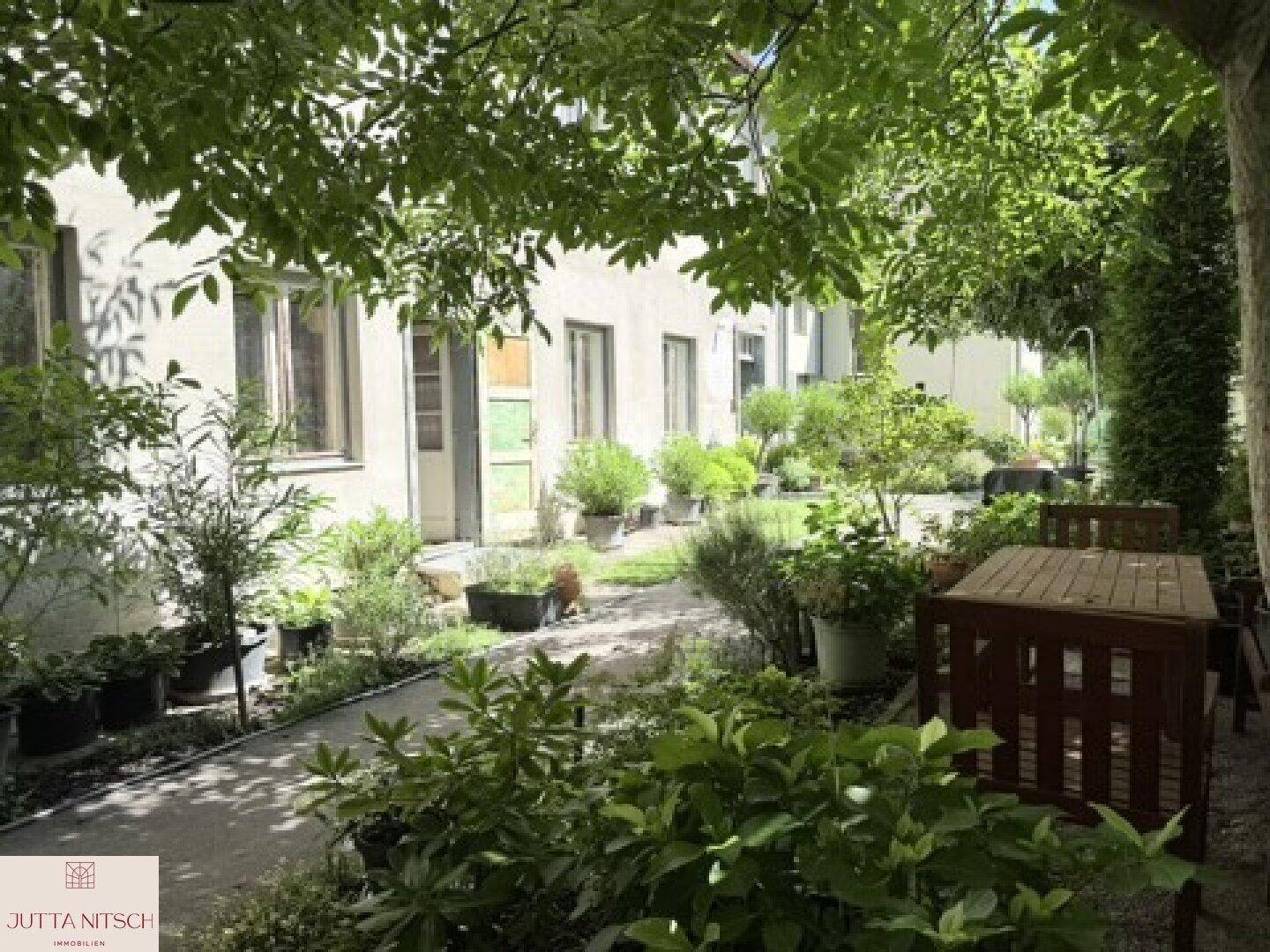 Schattiger Gartenbereich mit einem Holztisch und Stühlen, umgeben von üppigem Grün und Topfpflanzen.