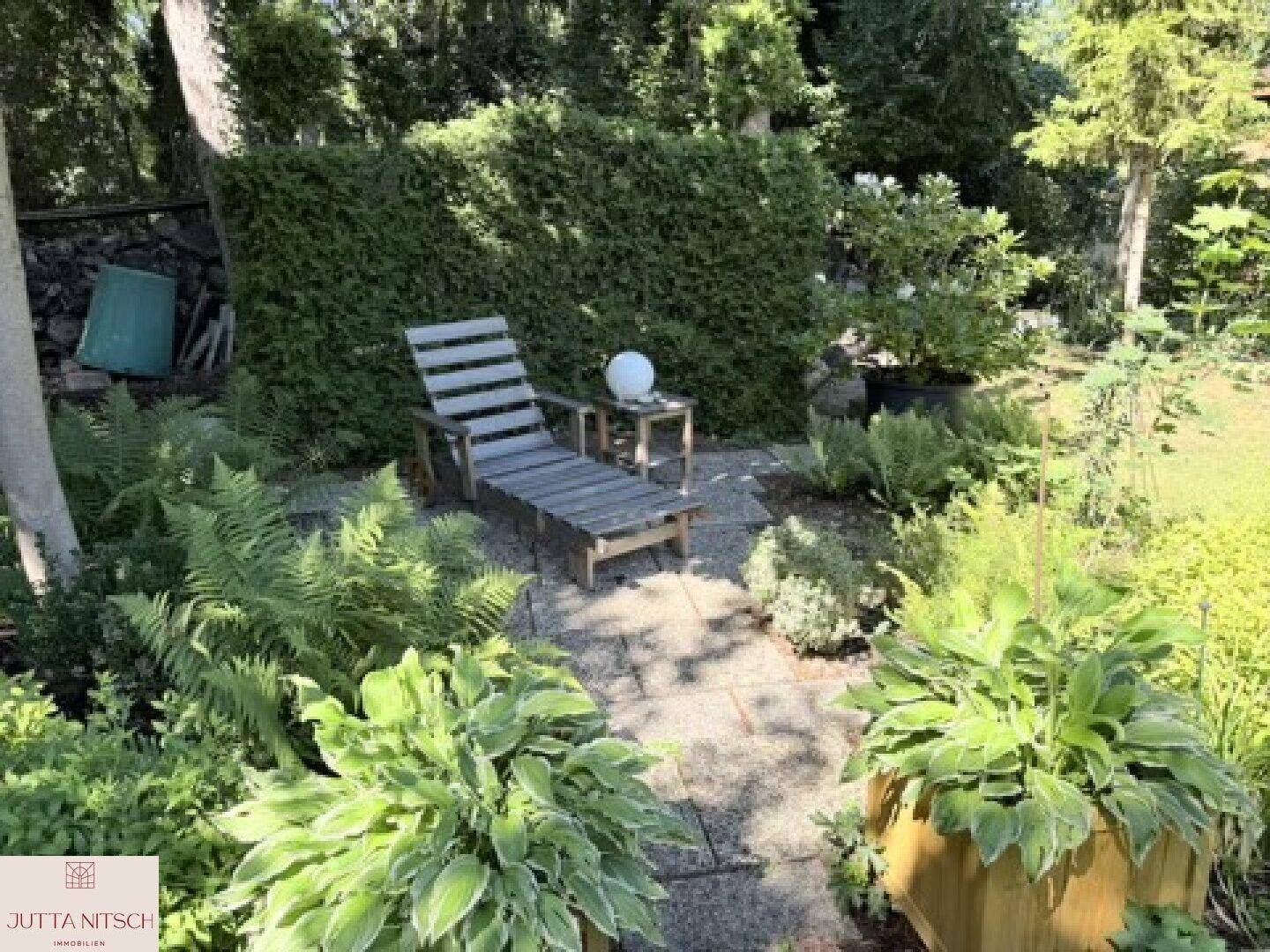 Ruhiger Gartenbereich mit einer Holzliege und einem Beistelltisch, umgeben von Farnen und Sträuchern.