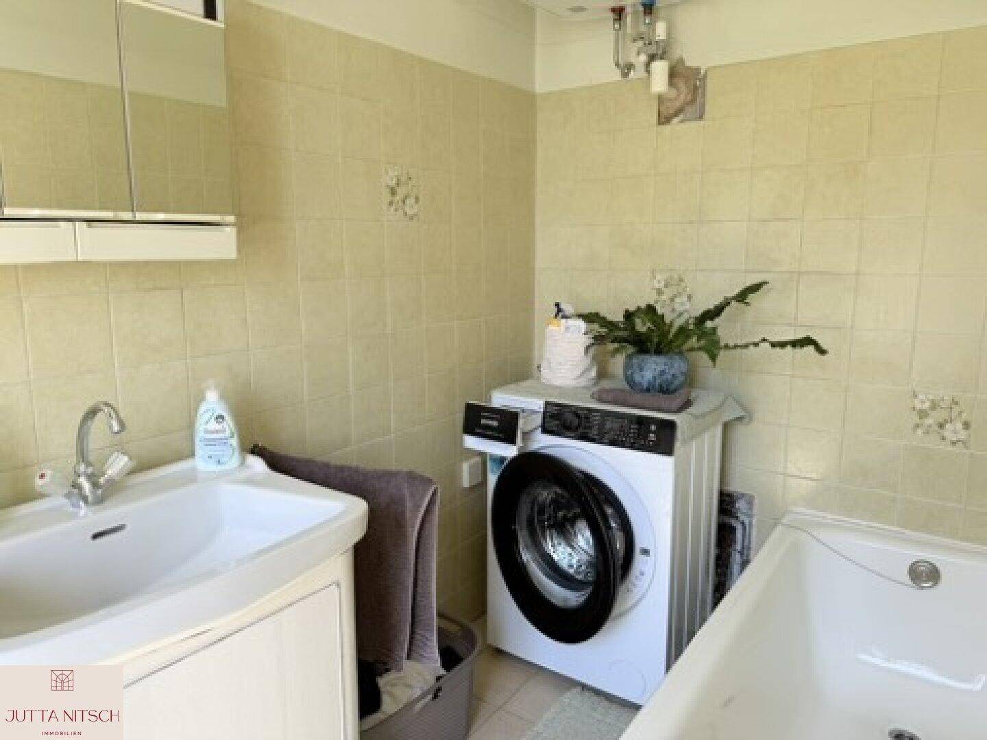Badezimmer mit beigefarbenen Fliesen, Waschbecken, Badewanne und einer Waschmaschine, die den Raum praktisch ergänzt.