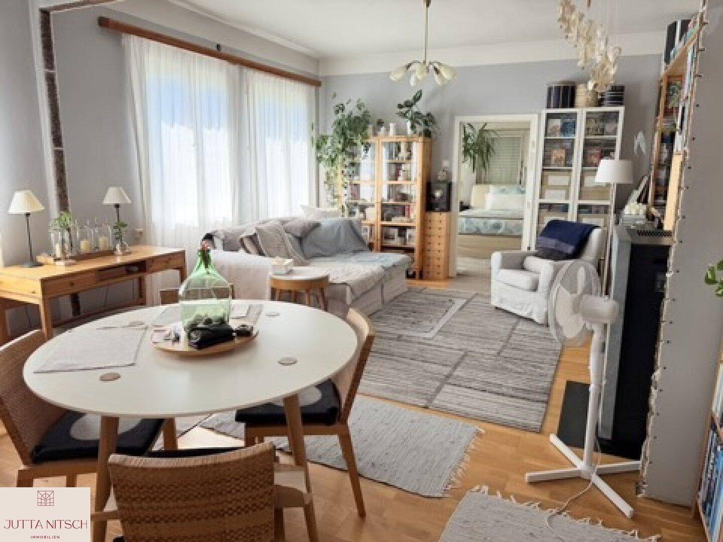 Gemütlicher Wohn- und Essbereich mit Holzboden, großem Fenster und verschiedenen Möbeln in hellen Farbtönen.