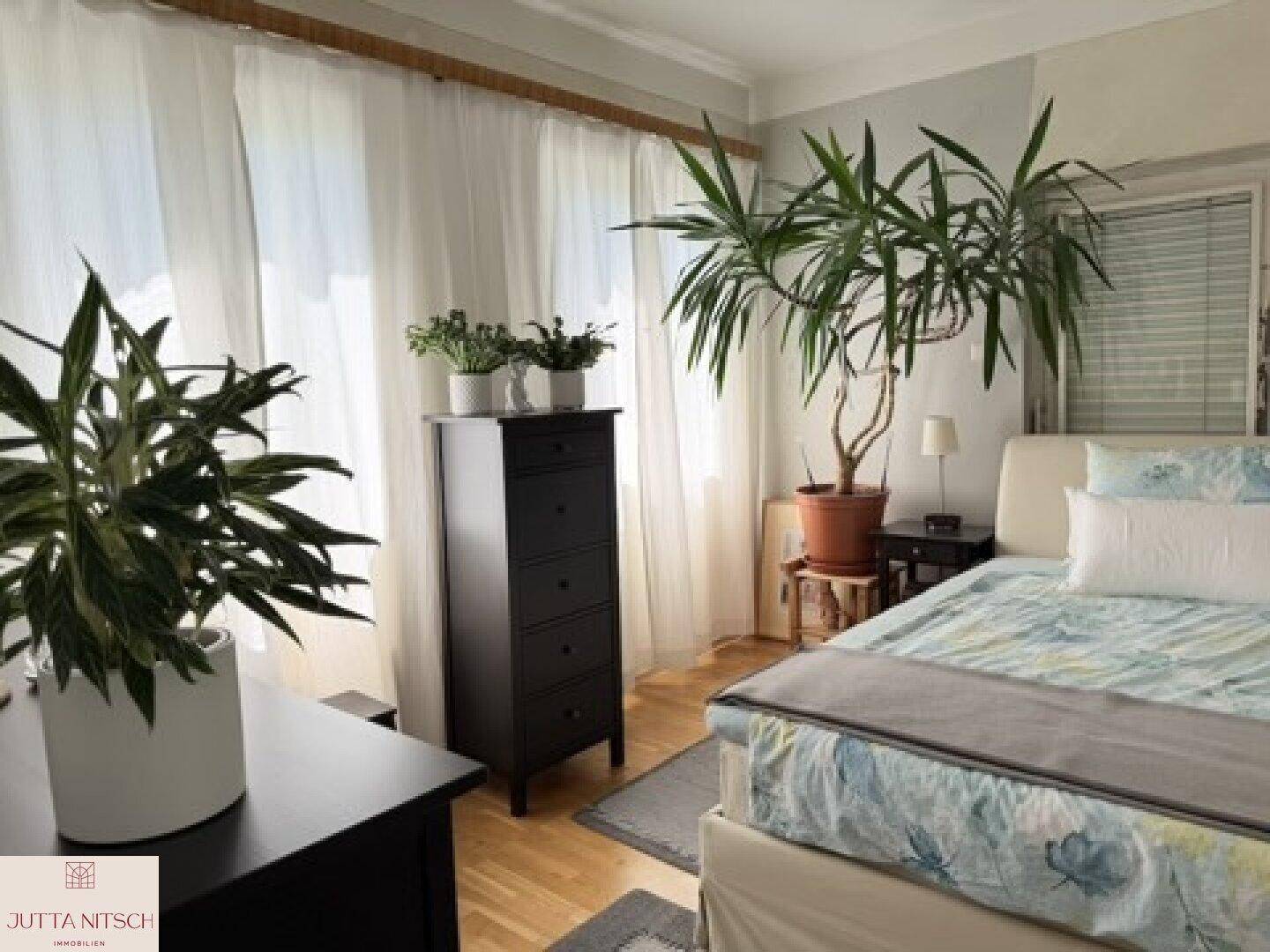 Helles Schlafzimmer mit Holzboden, großem Fenster, einem Bett und mehreren Grünpflanzen, die für eine frische Atmosphäre sorgen.