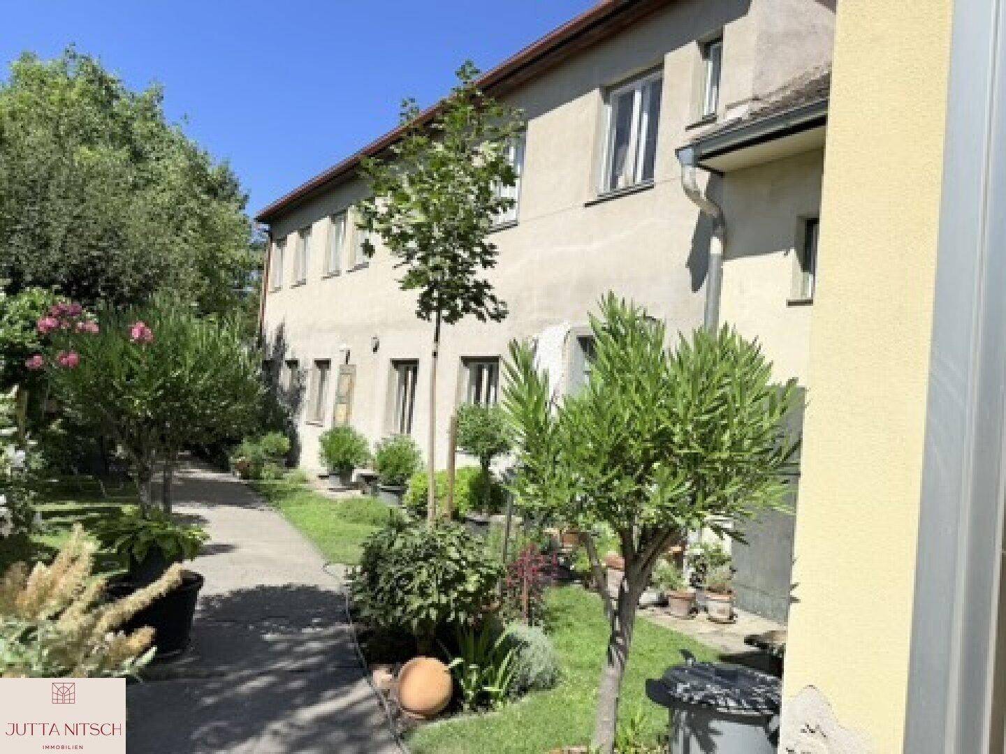 Seitenansicht des Gebäudes mit mehreren Fenstern und einem grünen Gartenbereich mit verschiedenen Pflanzen und Bäumen.