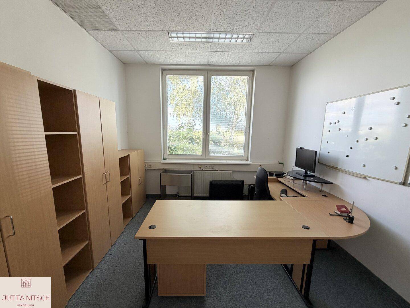 Funktionales Büro mit Schreibtisch, Bürostuhl, Regalen und einem Fenster mit Blick ins Grüne.