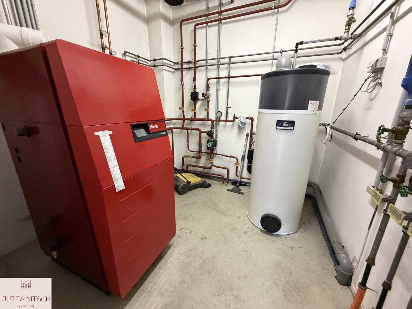 Technikraum mit einem großen roten Heizkessel und einem Warmwasserspeicher sowie sichtbaren Rohrleitungen.