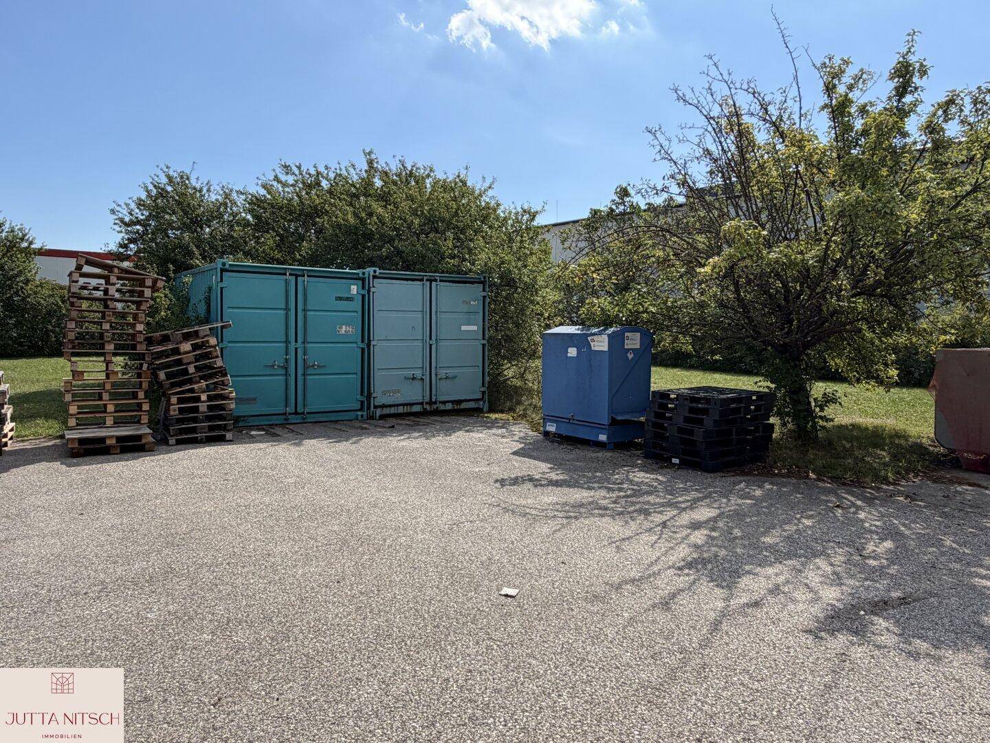 Außenbereich mit mehreren Lagercontainern und gestapelten Paletten, umgeben von Bäumen und Grasflächen.