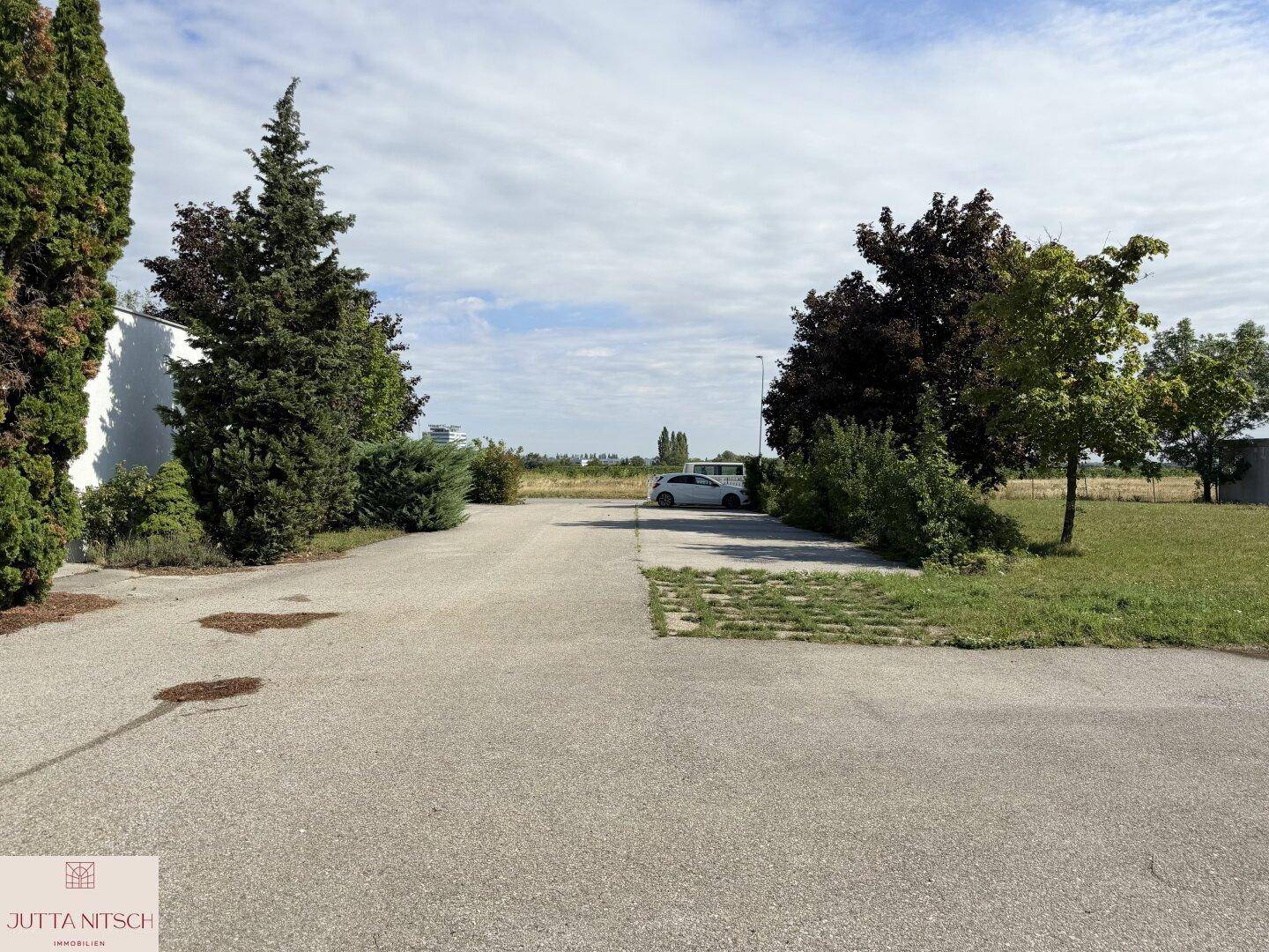 Außenbereich mit asphaltiertem Parkplatz, umgeben von Bäumen und einer weiten Landschaft im Hintergrund.