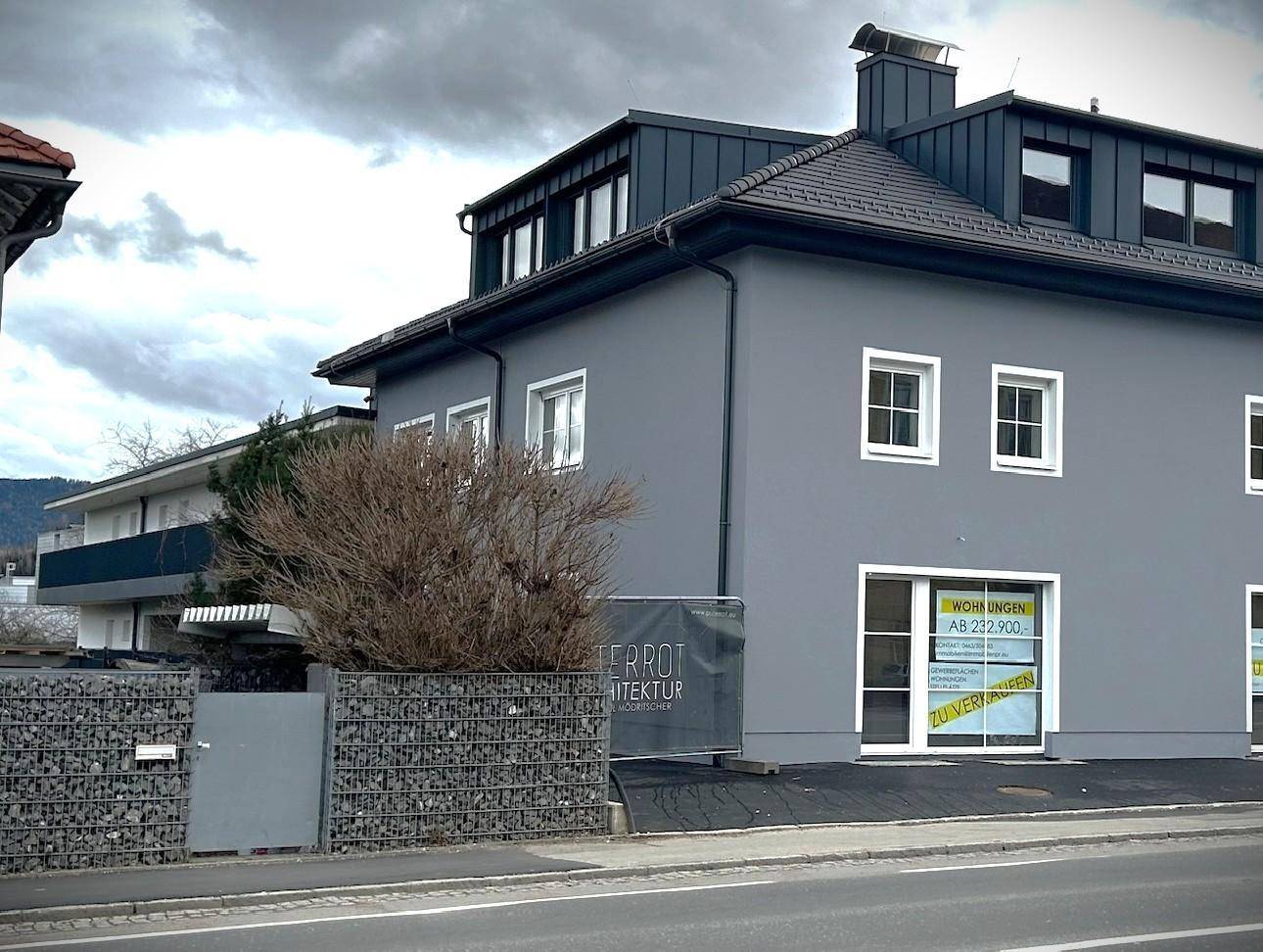 Ansicht des grauen Neubaus von der Straßenseite, mit einer Gabionenmauer im Vordergrund unter bewölktem Himmel.