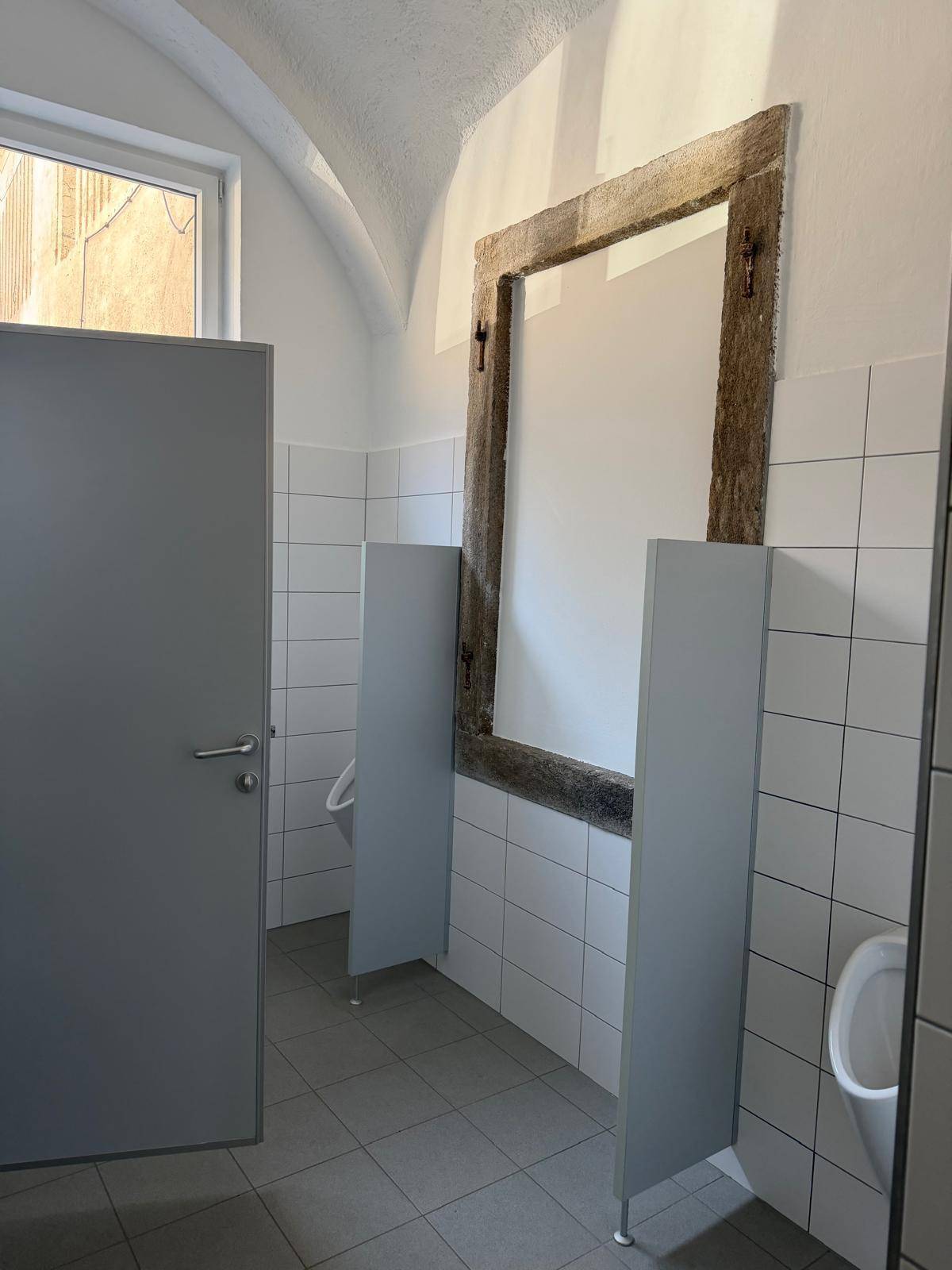 Modernes Badezimmer mit weißen Fliesen, grauen Trennwänden und Urinalen.