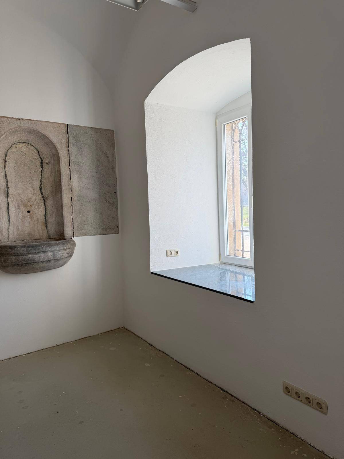 Ecke eines Raumes mit einem hohen Fenster und einer dekorativen Steininstallation an der Wand.