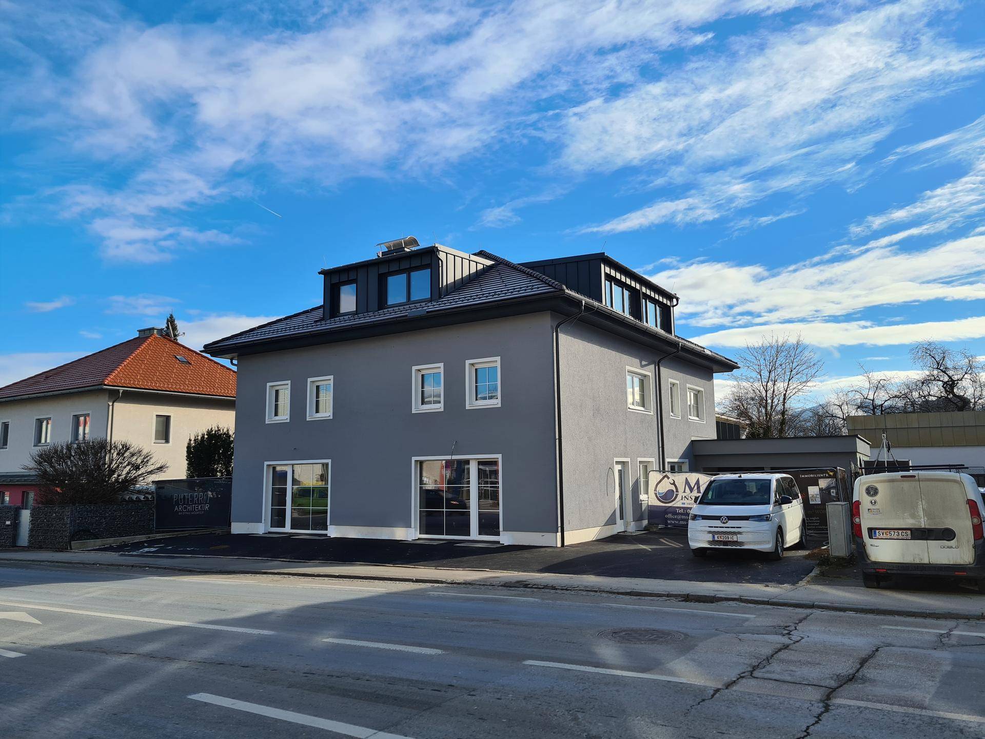 Gesamtansicht des modernen grauen Gebäudes von der Straße aus, mit geparkten Lieferwagen davor unter blauem Himmel.