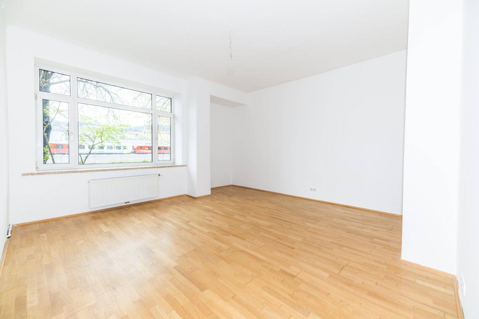 Helles, leeres Zimmer mit hellem Parkettboden, weißen Wänden und großem Fenster.