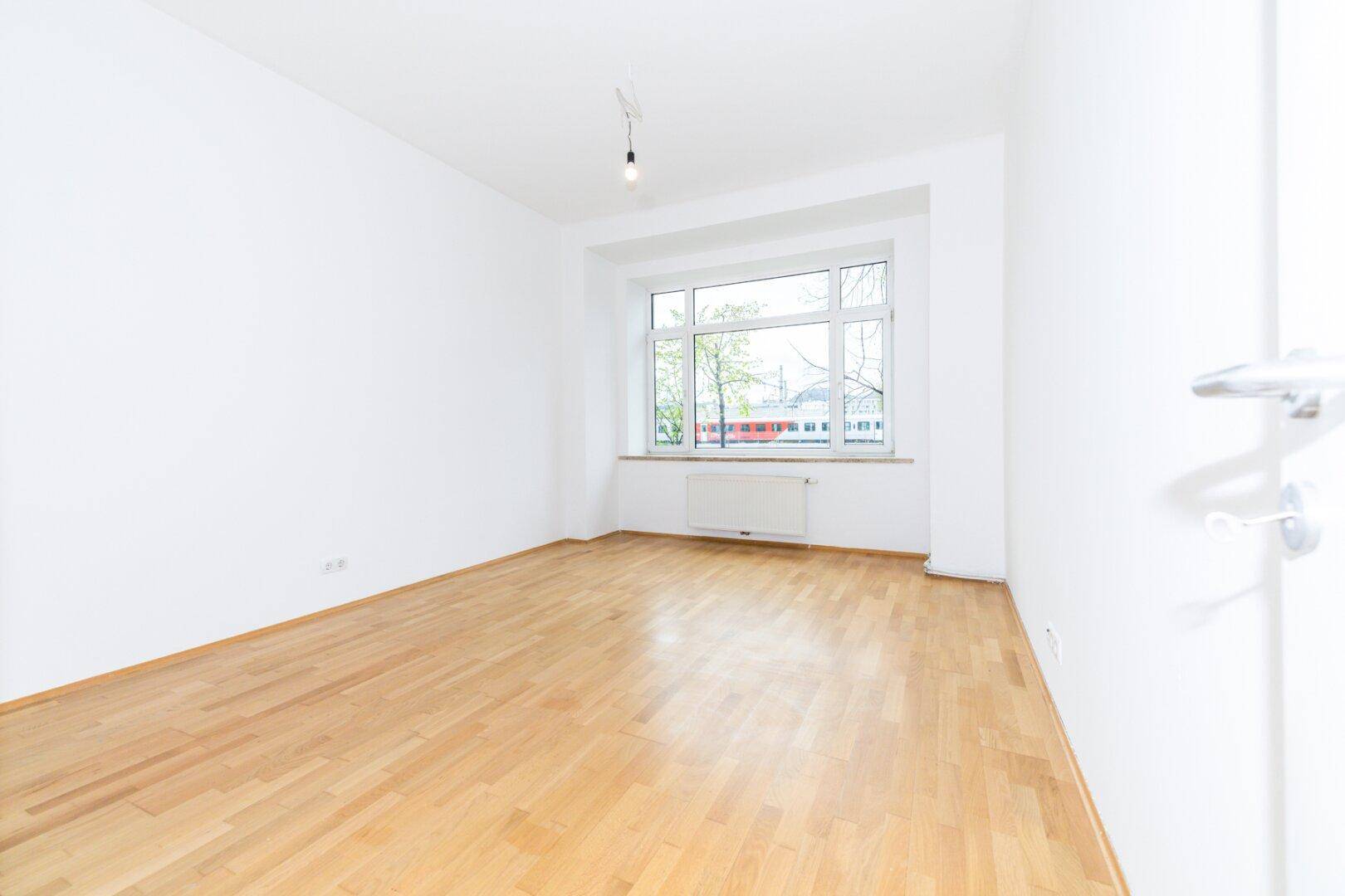 Geräumiges, leeres Zimmer mit hellem Parkettboden und großem Fenster für viel Tageslicht.