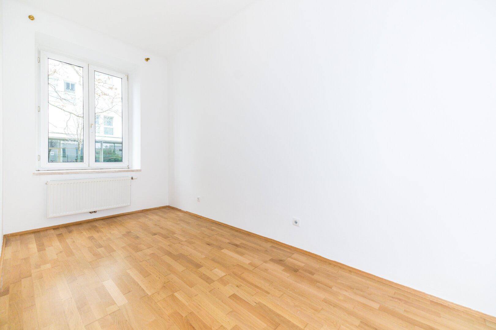 Helles, leeres Zimmer mit Parkettboden und einem Fenster, das den Blick ins Grüne freigibt.