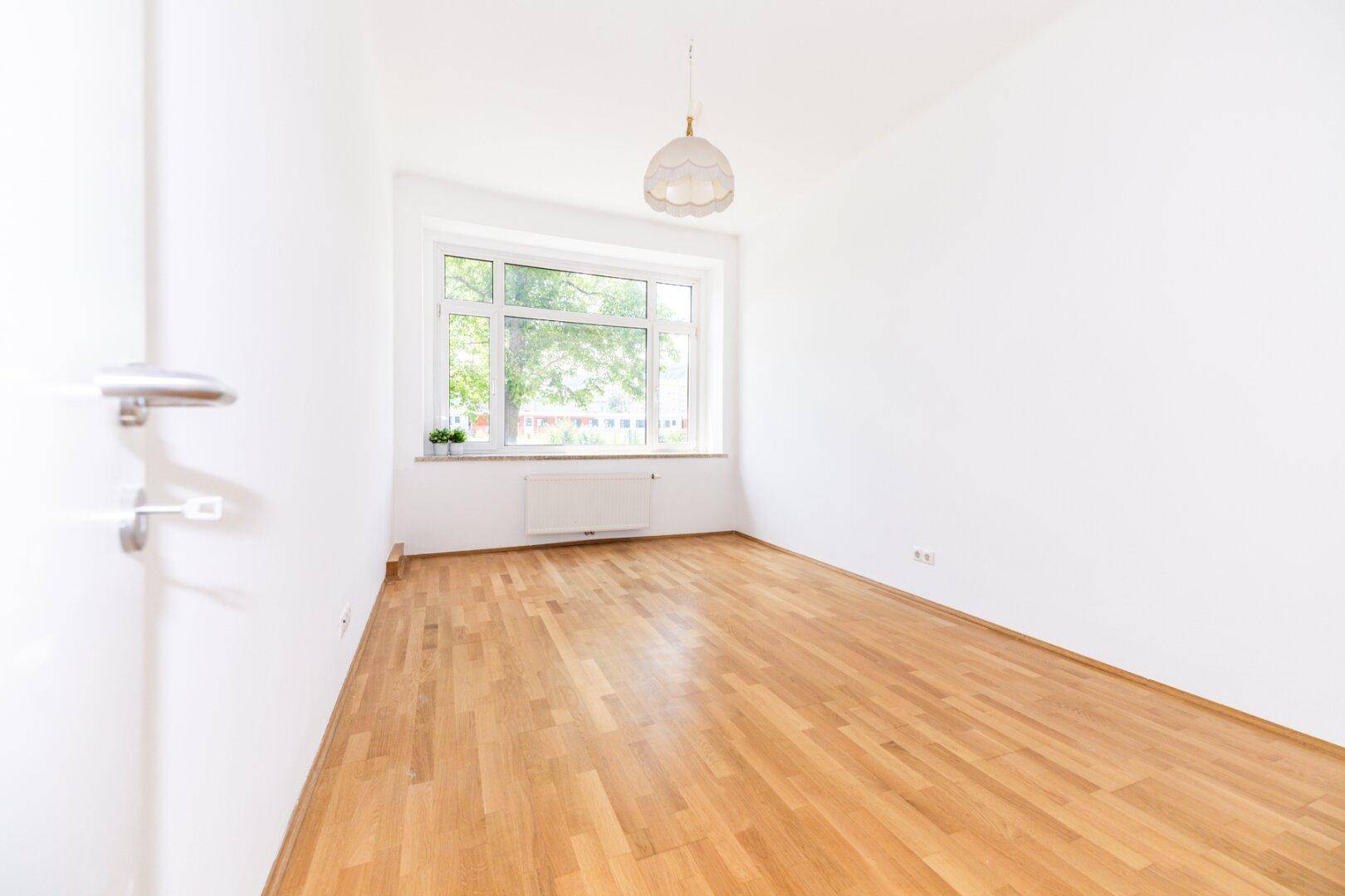 Leeres Zimmer mit hellem Parkettboden, weißen Wänden und einem Fenster mit Heizkörper.