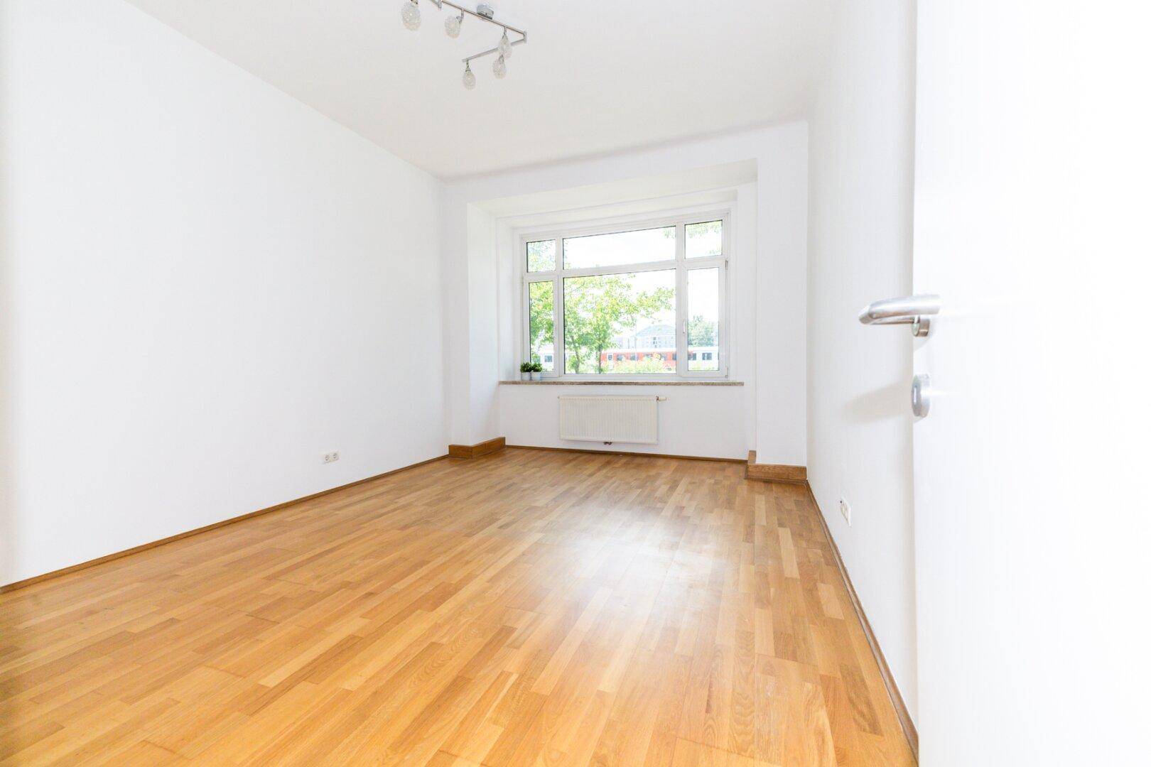 Helles, geräumiges Zimmer mit Parkettboden, weißen Wänden und einem großen Fenster.