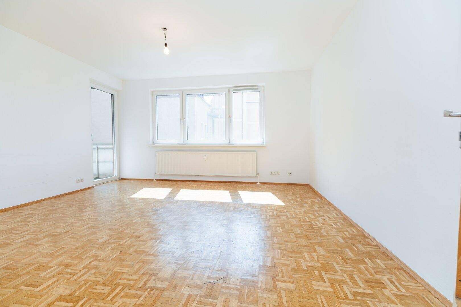 Geräumiges Wohnzimmer mit Parkettboden, großen Fenstern und Zugang zum Balkon.