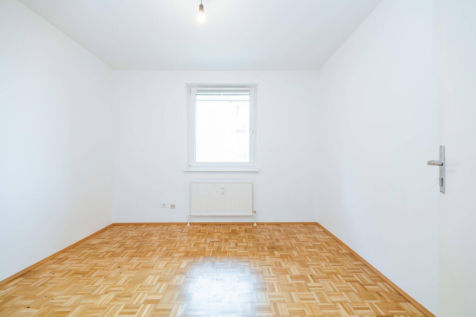 Leeres Zimmer mit Parkettboden und einem Fenster, das viel Tageslicht hereinlässt.