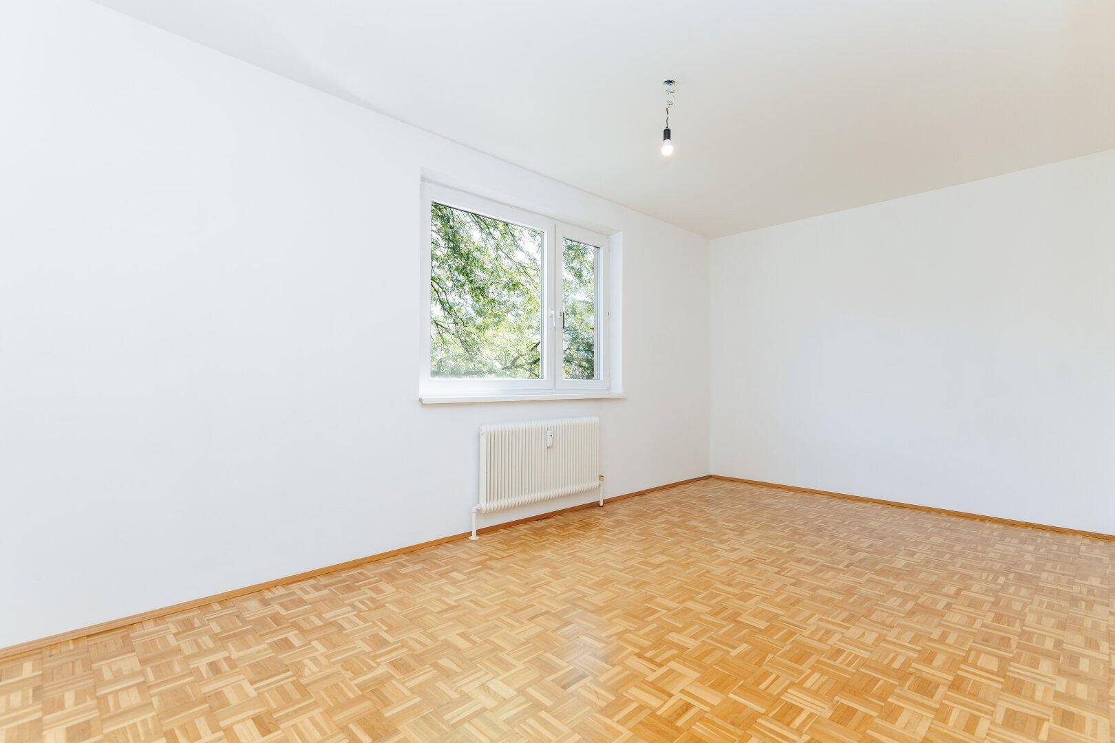 Helles, leeres Zimmer mit Parkettboden und großem Fenster, das viel Tageslicht hereinlässt.