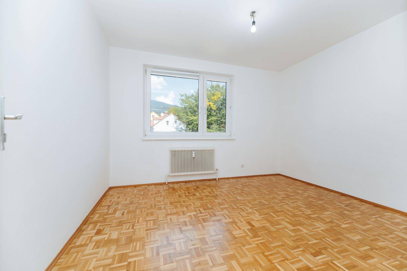 Leeres Schlafzimmer mit Parkettboden und Fenster, das einen Blick auf die Umgebung bietet.