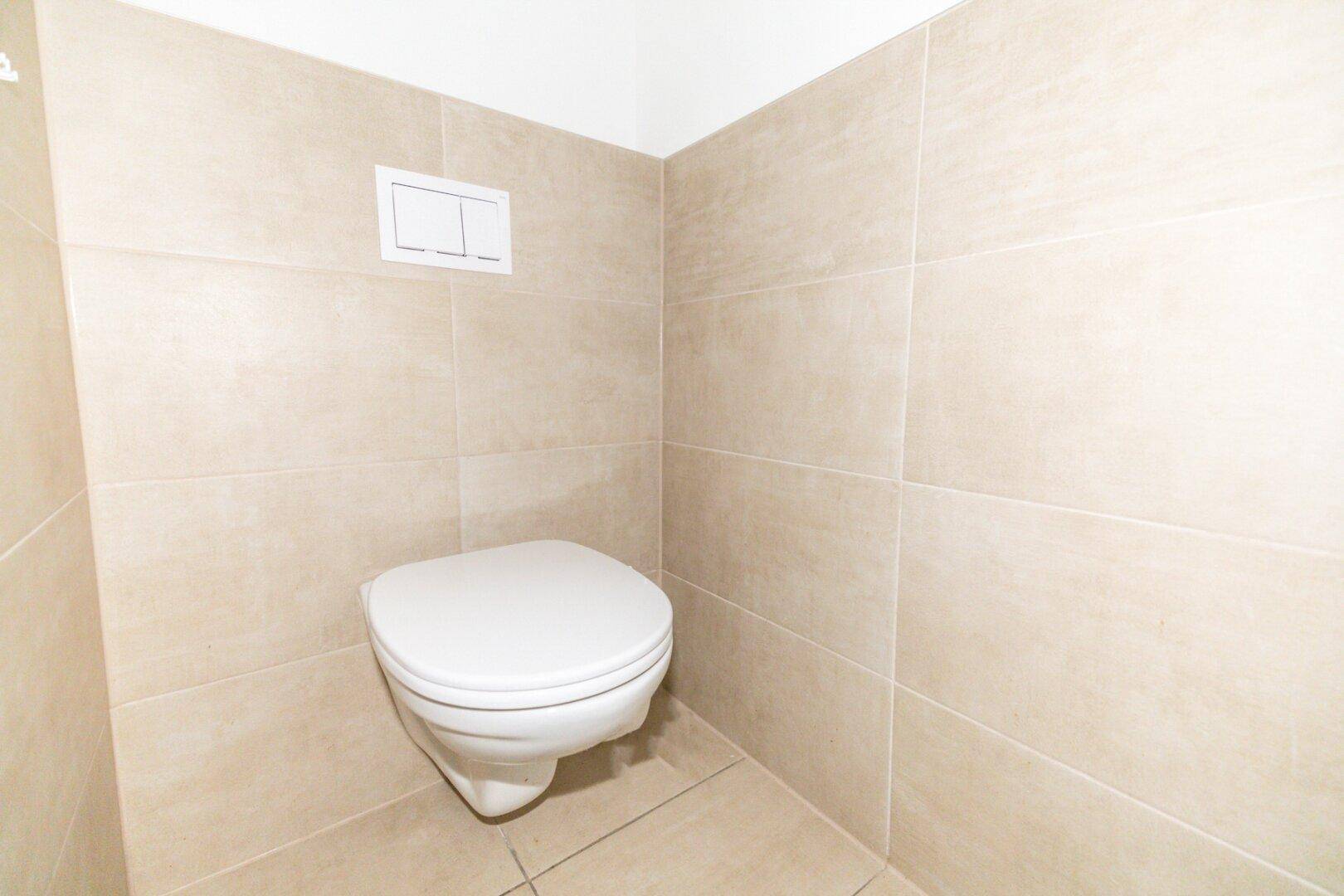 Wandhängendes WC mit beigefarbenen Fliesen an den Wänden und auf dem Boden.