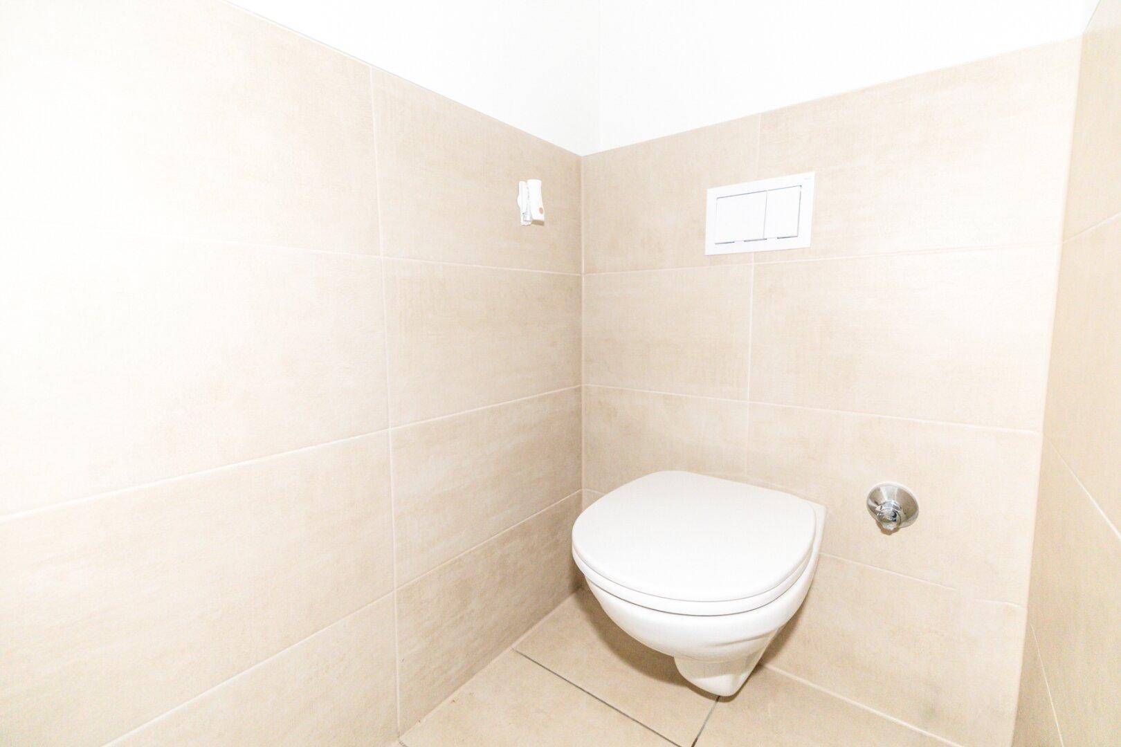 Wandhängendes WC in einem gefliesten Raum mit beigefarbenen Wand- und Bodenfliesen.