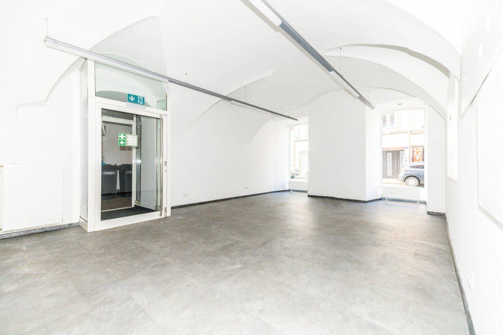 Geräumiger Eingangsbereich mit Gewölbedecken, Betonboden und großen Glastüren zur Straße.