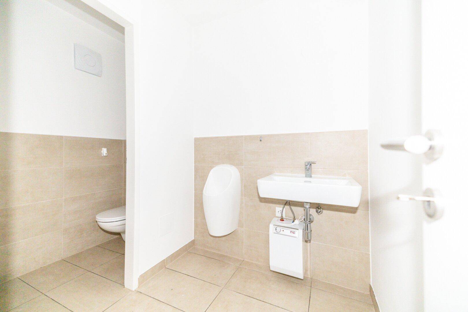 Modernes WC mit Urinal, Waschbecken und beigefarbenen Fliesen an Wänden und Boden.